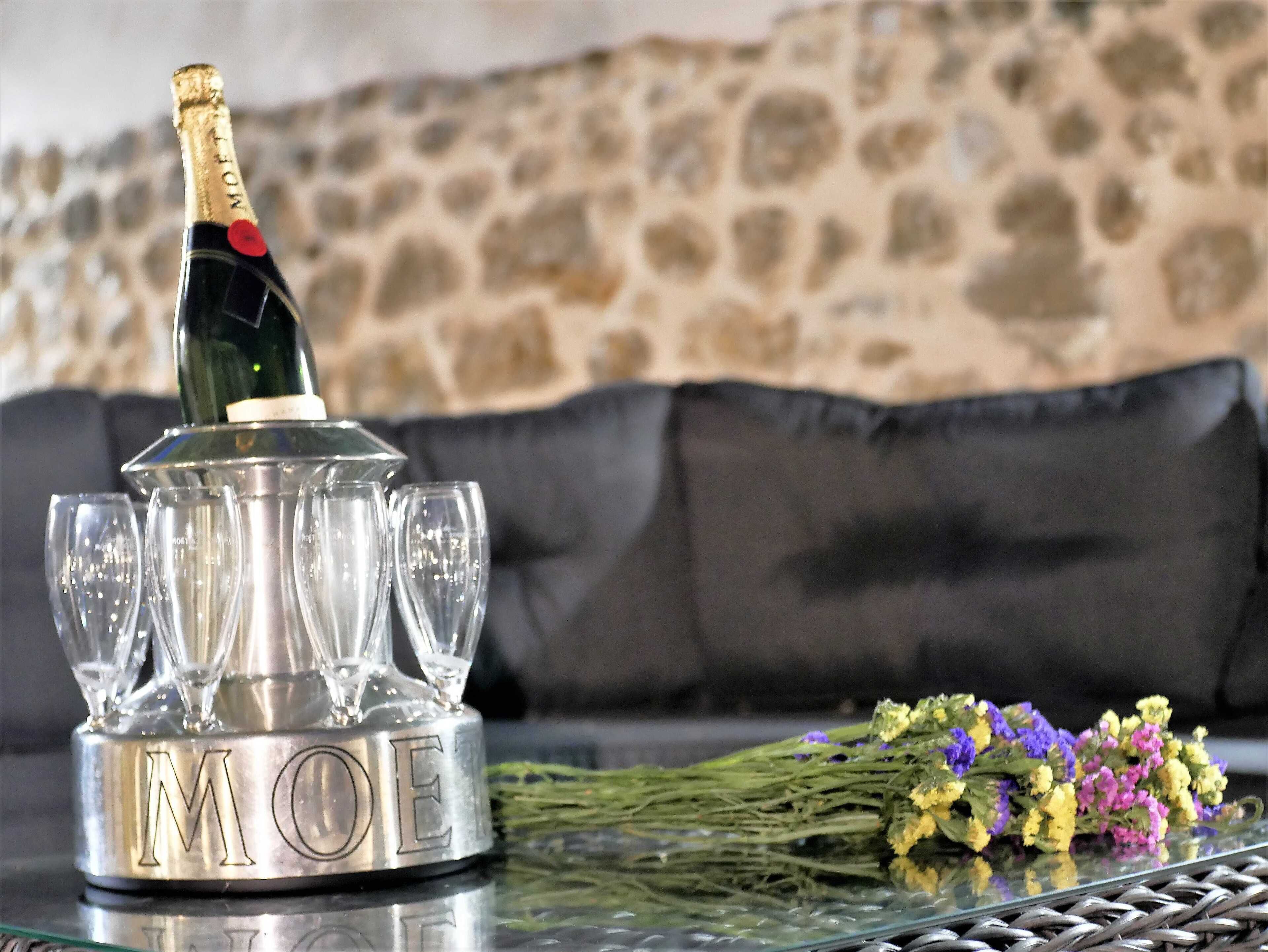 A champagne bottle on a metallic stand with glasses, a bouquet of flowers, and a stone wall in the background.