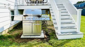 A stainless steel grill with four burners and a side cabinet, located on a concrete pad in front of a white house with a balcony and a staircase. - Newly renovated! 5 bedroom 2 bath home woth AC and walk to water (Narragansett)