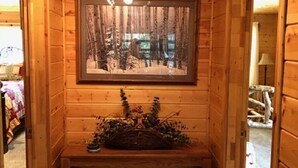 Interior de una cabaña de madera con un cuadro enmarcado de un bosque nevado en la pared, un banco con una cesta y una lámpara en el techo.