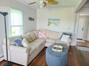 A living room with a beige sectional sofa, a blue ottoman, a floor lamp, and a ceiling fan.