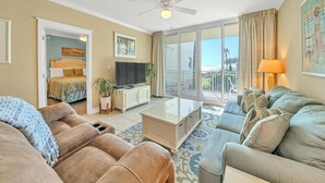 A spacious living room with a sofa set, a glass coffee table, a television, and a balcony with a view.