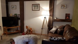 Een woonkamer met een televisie op een houten standaard, een driepootlamp, een bruine leren bank met geknoopde kussens, en een rode stoel met een witte bont plaid.