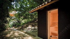 Ein abgelegener Außenbereich mit einem Kiesweg, der zu einer Holzhütte mit Glastür führt, umgeben von üppigem Grün und zwei Korbstühlen.