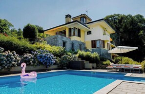 Uma casa moderna de dois andares com piscina, rodeada por vegetação exuberante e um boi flamingo flutuante.