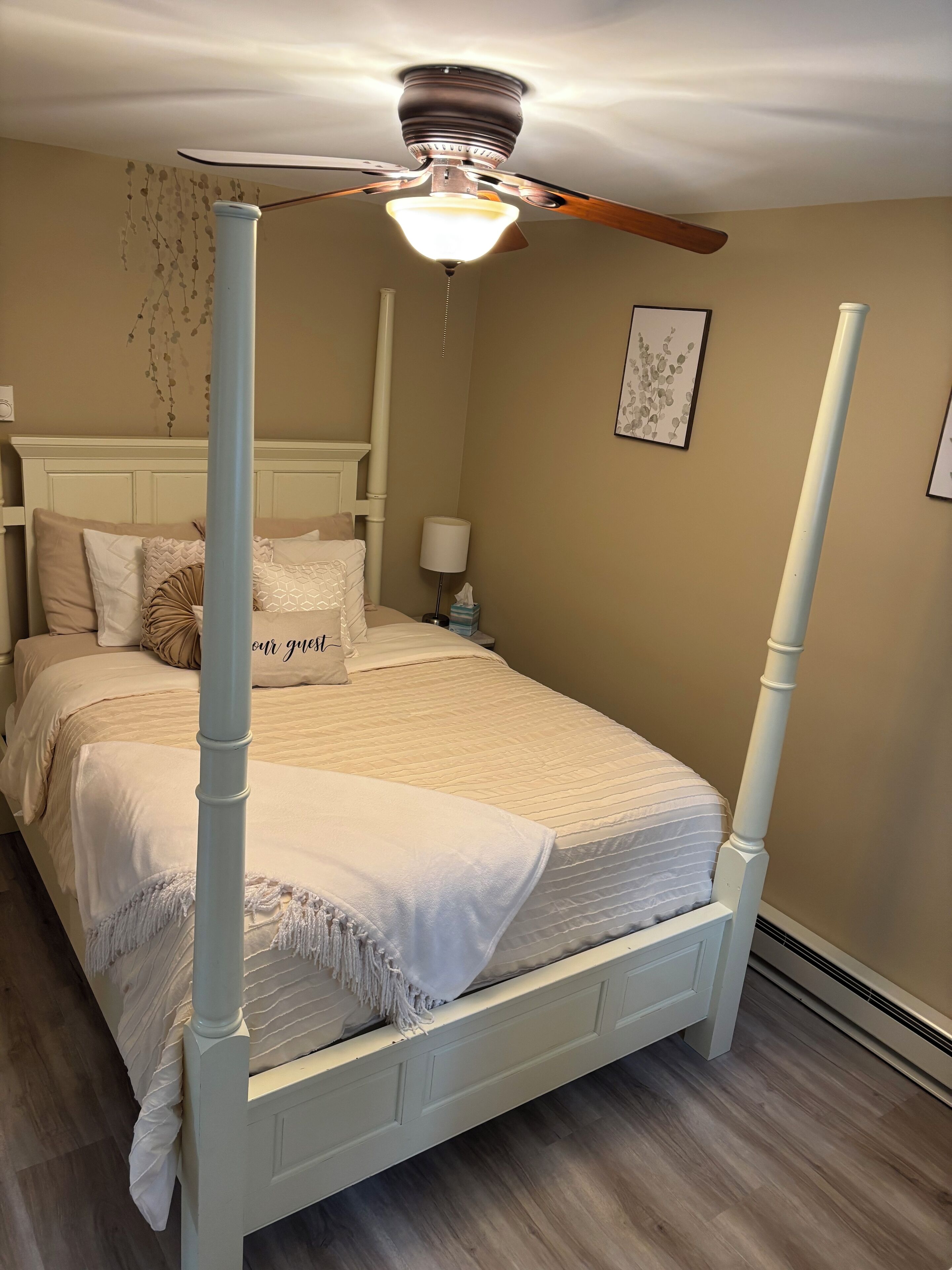 A four-poster bed with a canopy, a bedside table with a lamp, and a wall-mounted picture.
