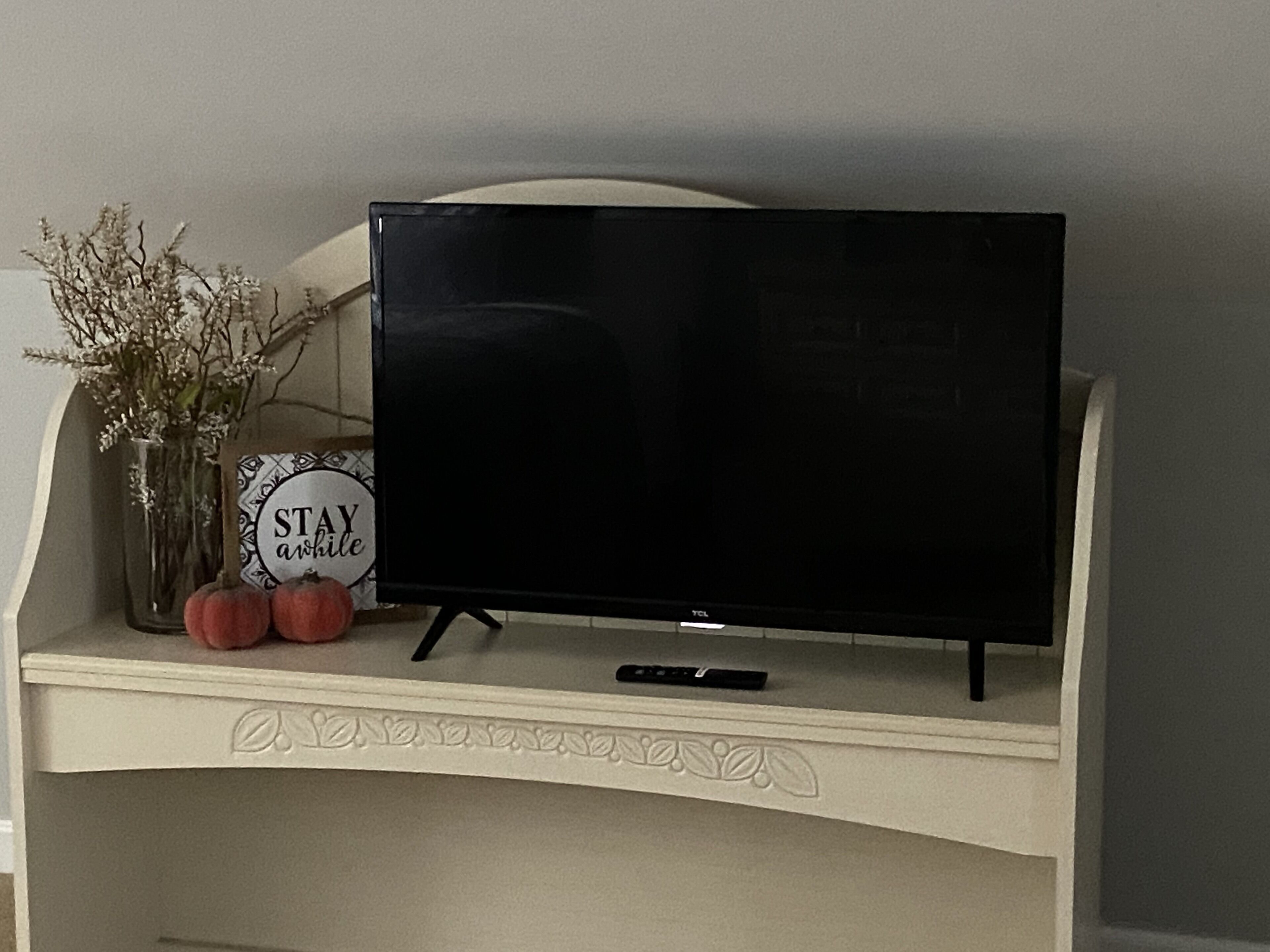 Un televisore su uno scaffale bianco, decorato con foglie ornamentali, un vaso con fiori secchi e un cartello con su scritto “STAY awhile”.