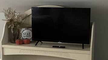 Un televisore su uno scaffale bianco, decorato con foglie ornamentali, un vaso con fiori secchi e un cartello con su scritto “STAY awhile”.