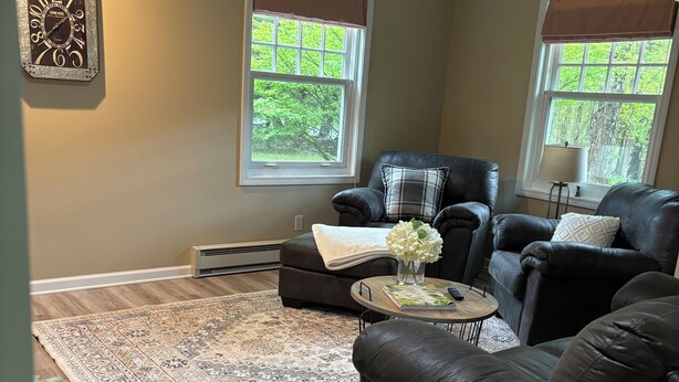A living room with a dark leather sofa, a coffee table, and a patterned rug. There are two windows with blinds, a ceiling fan, and a decorative clock on the wall. -  Come,Stay a While-WYNN Hosp.,Nexus Center,Cooperstown,Utica,Herkimer, Old Forge (Clayville)
