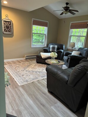 A living room with a dark leather sofa, a coffee table, and a patterned rug. There are two windows with blinds, a ceiling fan, and a decorative clock on the wall. -  Come,Stay a While-WYNN Hosp.,Nexus Center,Cooperstown,Utica,Herkimer, Old Forge (Clayville)