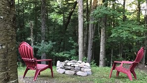 Two red Adirondack chairs facing a stone fire pit in a forested area. -  Come,Stay a While-WYNN Hosp.,Nexus Center,Cooperstown,Utica,Herkimer, Old Forge (Clayville)