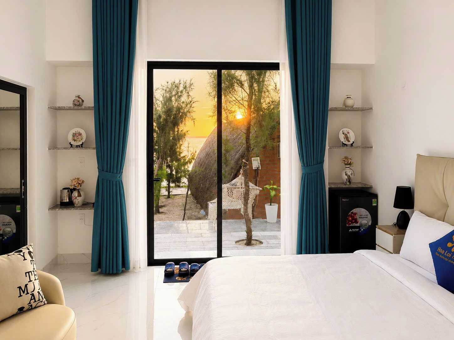 Habitación de hotel con cama, una silla, un escritorio y vistas a una playa al atardecer.