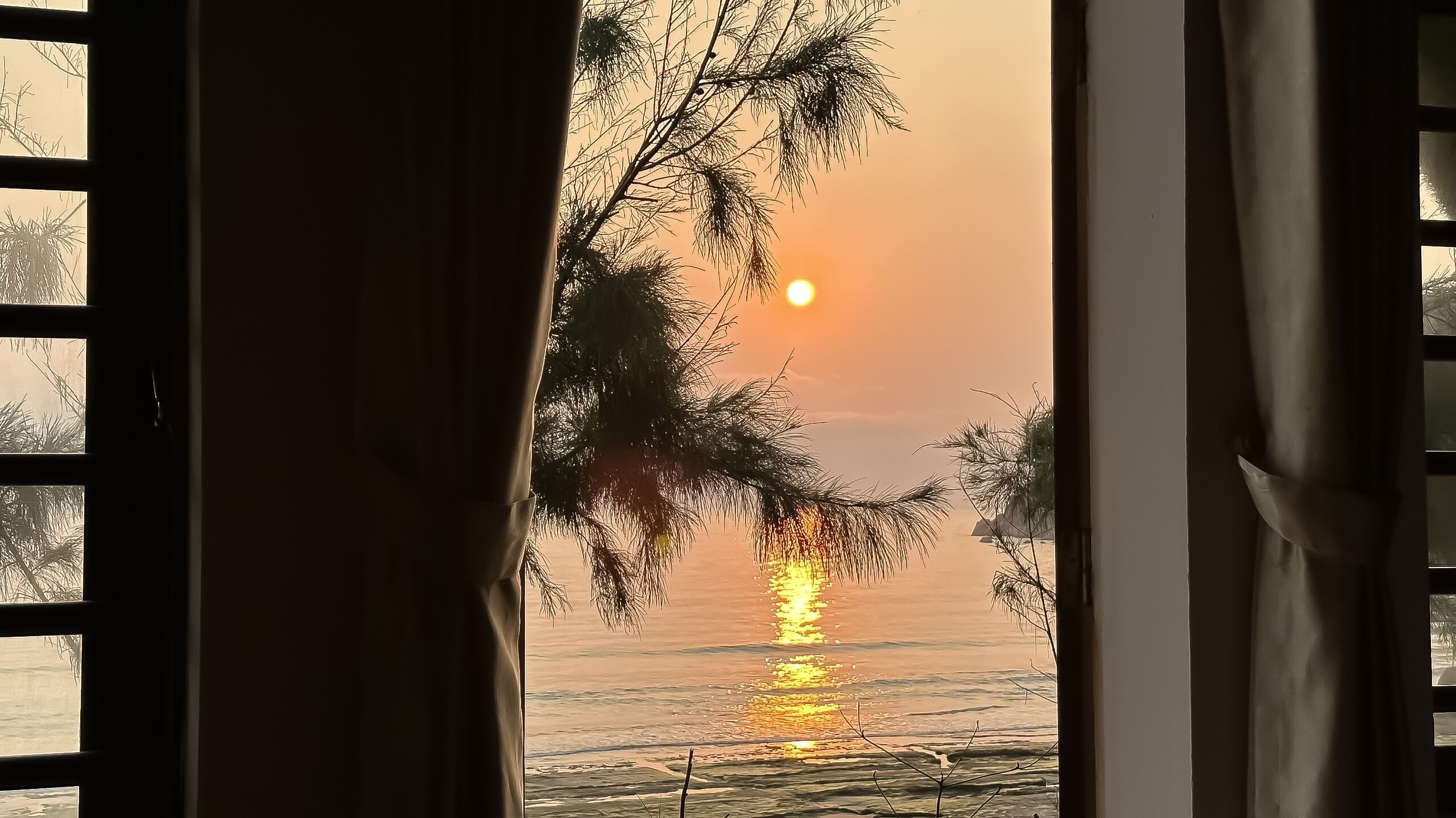A view through a window with curtains, featuring a sunset over a calm body of water with trees in the foreground.