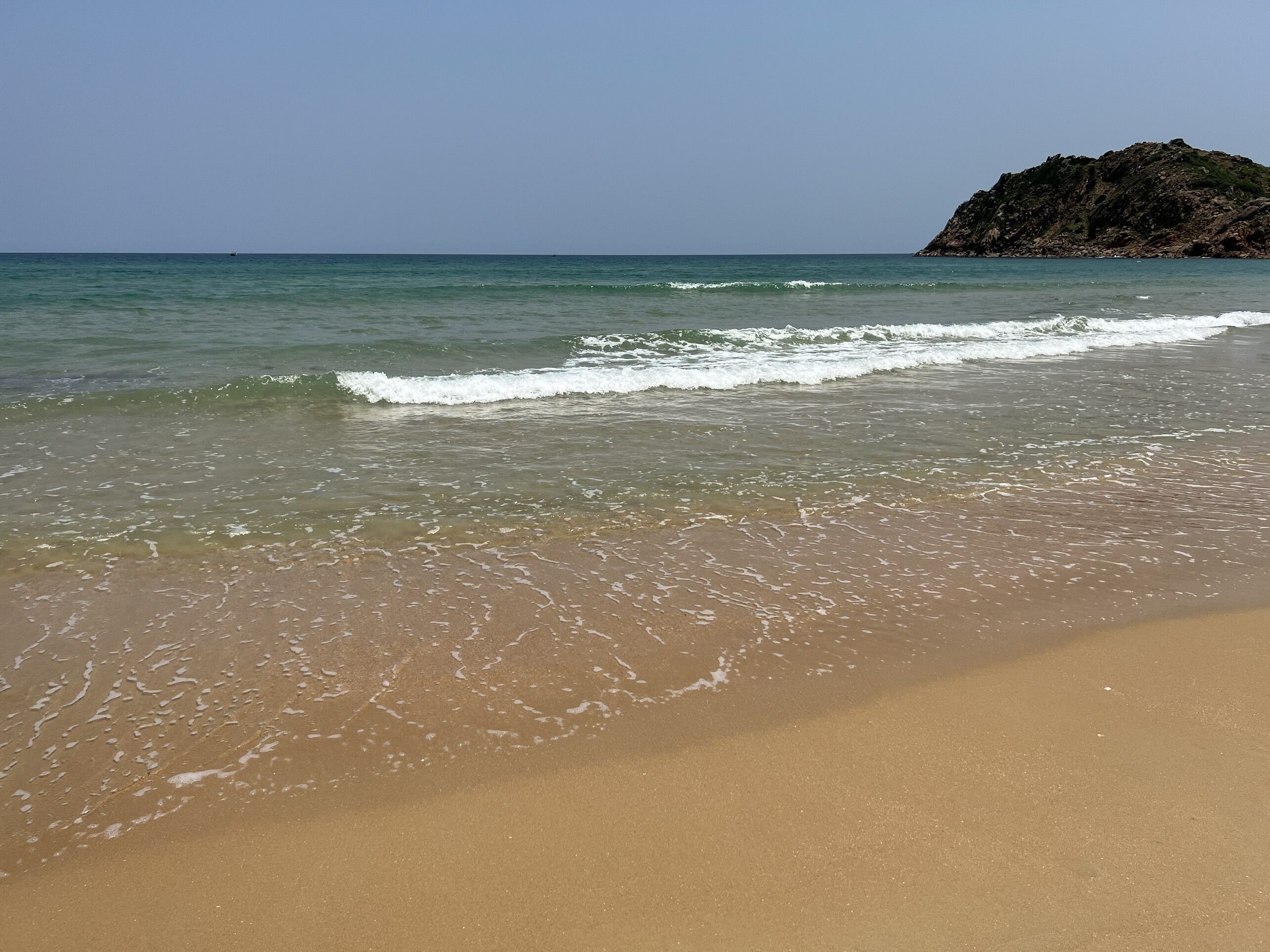 A beach with clear water, a sandy shore, and a rocky outcrop in the distance.