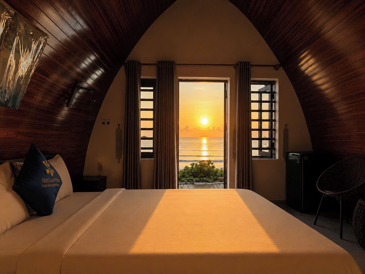 A bedroom with a large bed, a view of the sunset over the sea, and wooden walls.