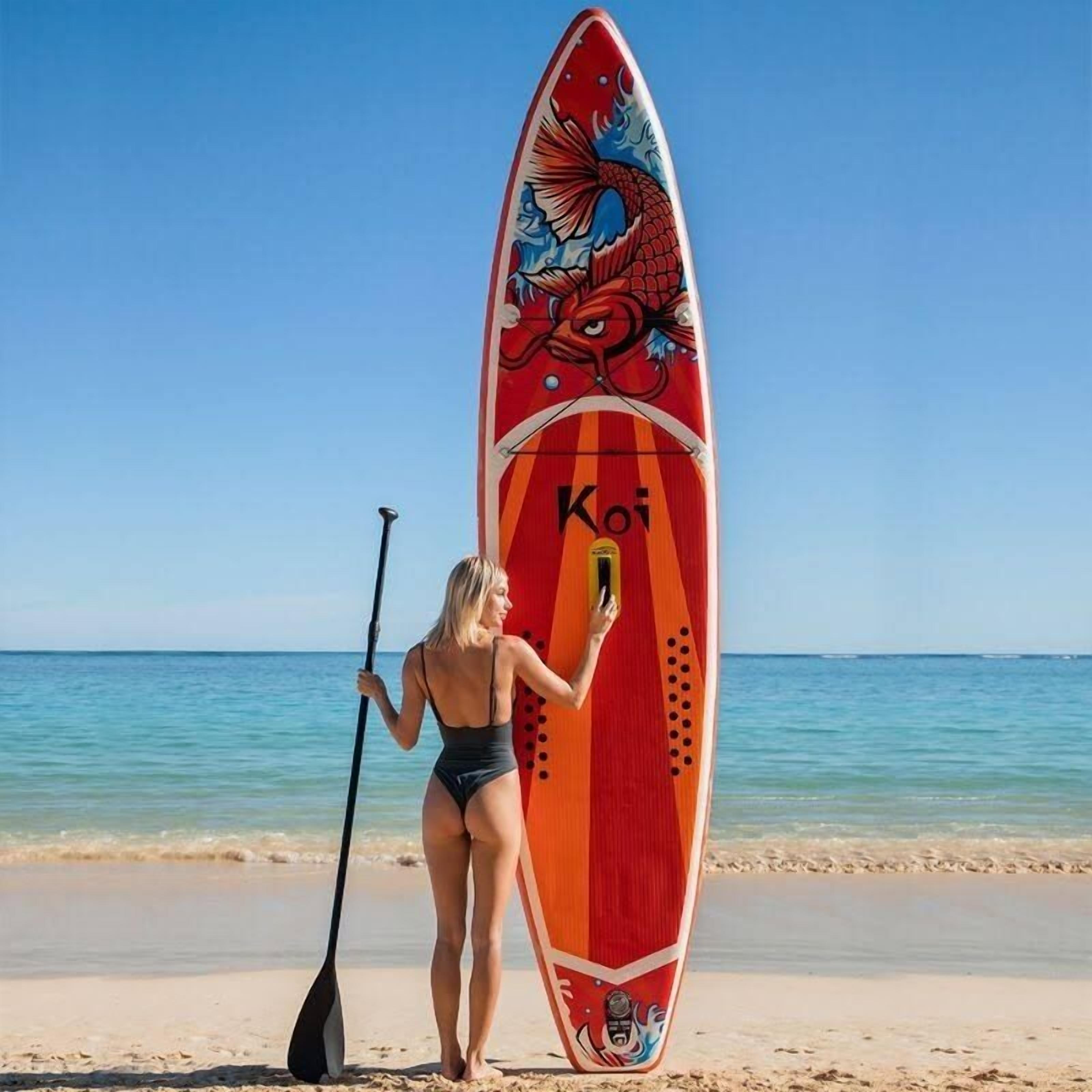 Una persona parada en la playa sosteniendo una tabla de remo con un diseño de dragón y la palabra "Koi".