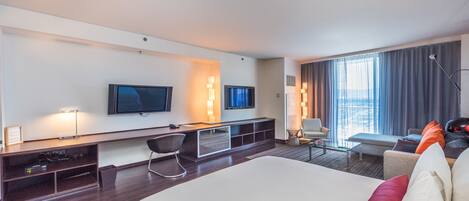 A modern hotel room with a large bed, a desk with a computer, a TV, and a seating area with a view of the city.