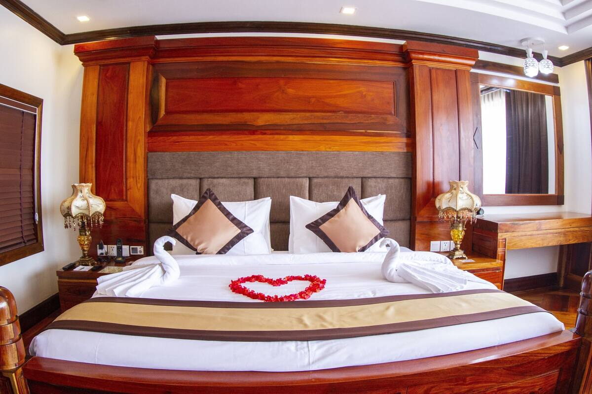 Una cama grande con decoración en forma de corazón, cabecera de madera y dos lámparas de noche.