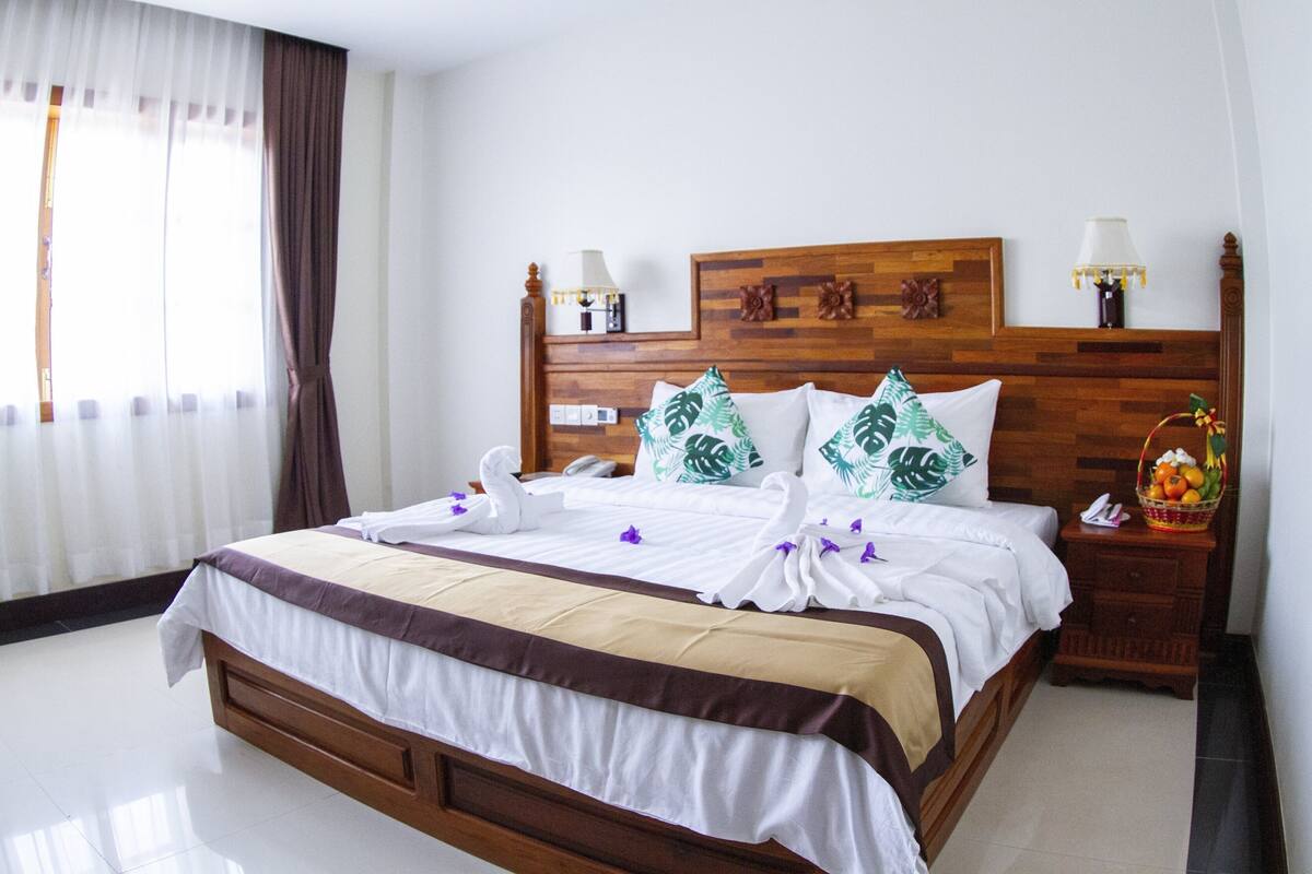Una cama bien hecha con cabecera de madera, ropa de cama blanca y almohadas decorativas. Hay una mesita de noche con una lámpara y una cesta de frutas.