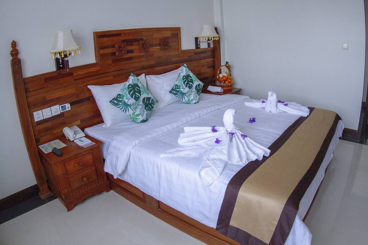 Una cama bien hecha con sábanas blancas, dos almohadas con estampado de hojas verdes y un corredor decorativo marrón. Hay dos lámparas de noche, una mesita de noche de madera con un teléfono y una pequeña mesa con una cesta de frutas y un objeto decorativo.