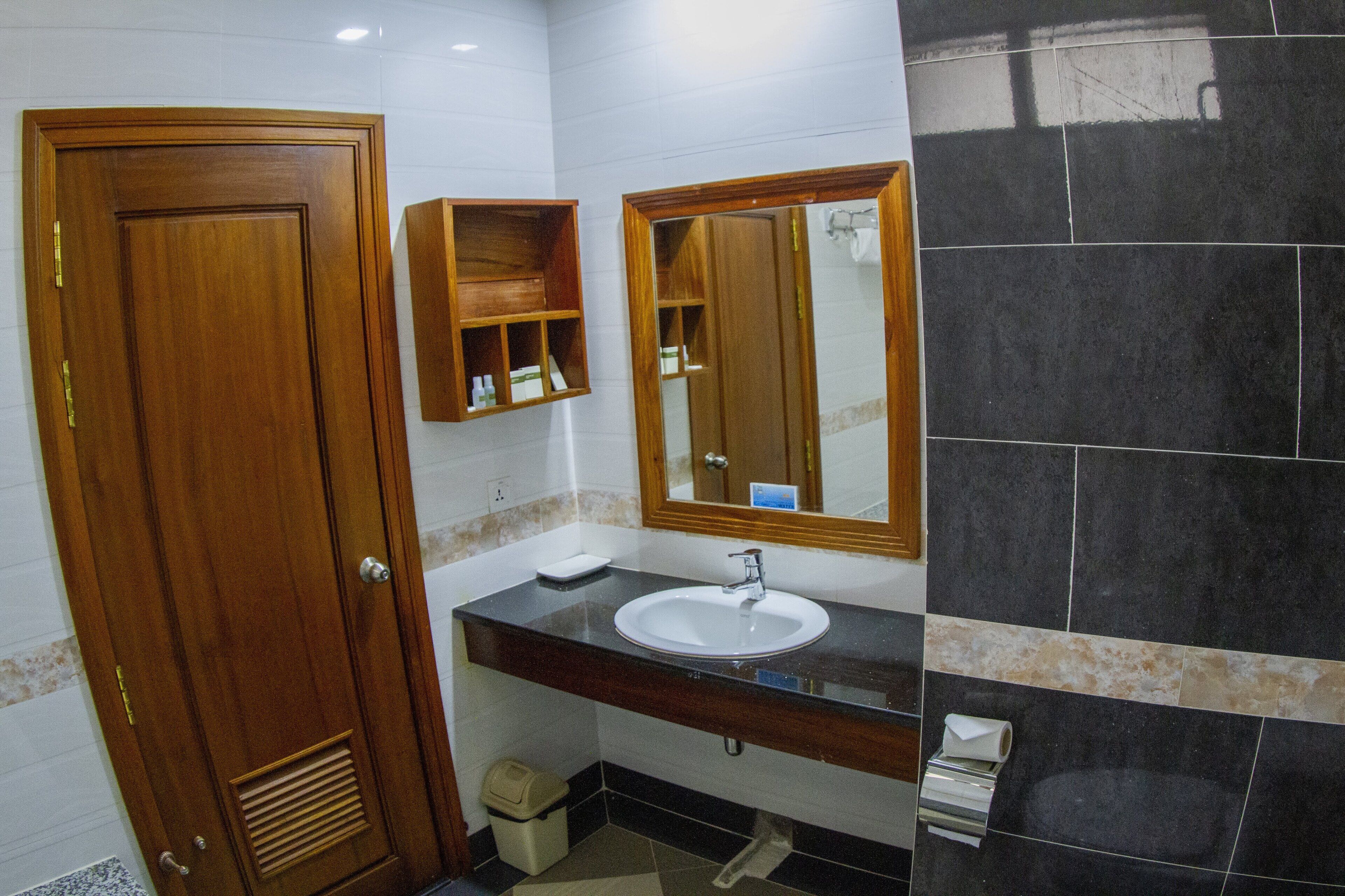 A bathroom with a wooden door, a white sink, a mirror, and a black tiled wall.