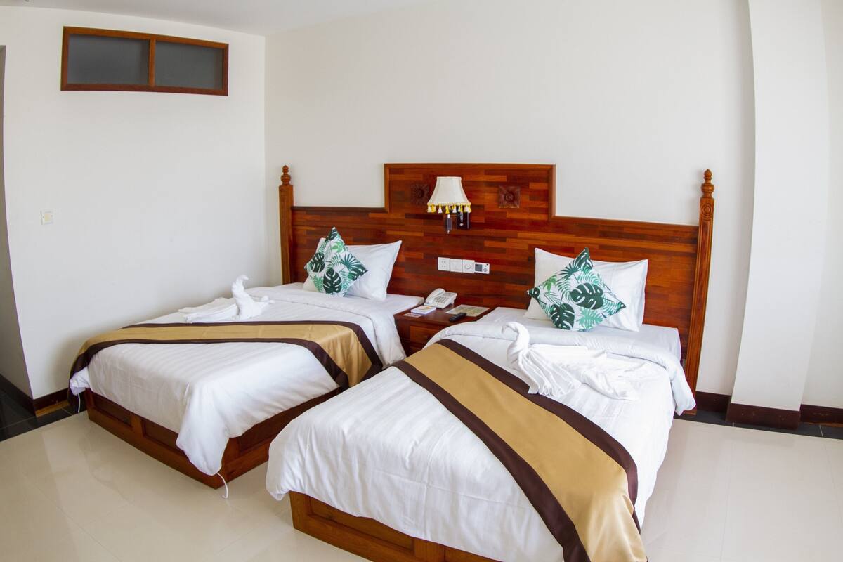 Habitación de hotel con dos camas, cabeceros de madera, ropa de cama blanca y una lámpara de pared.