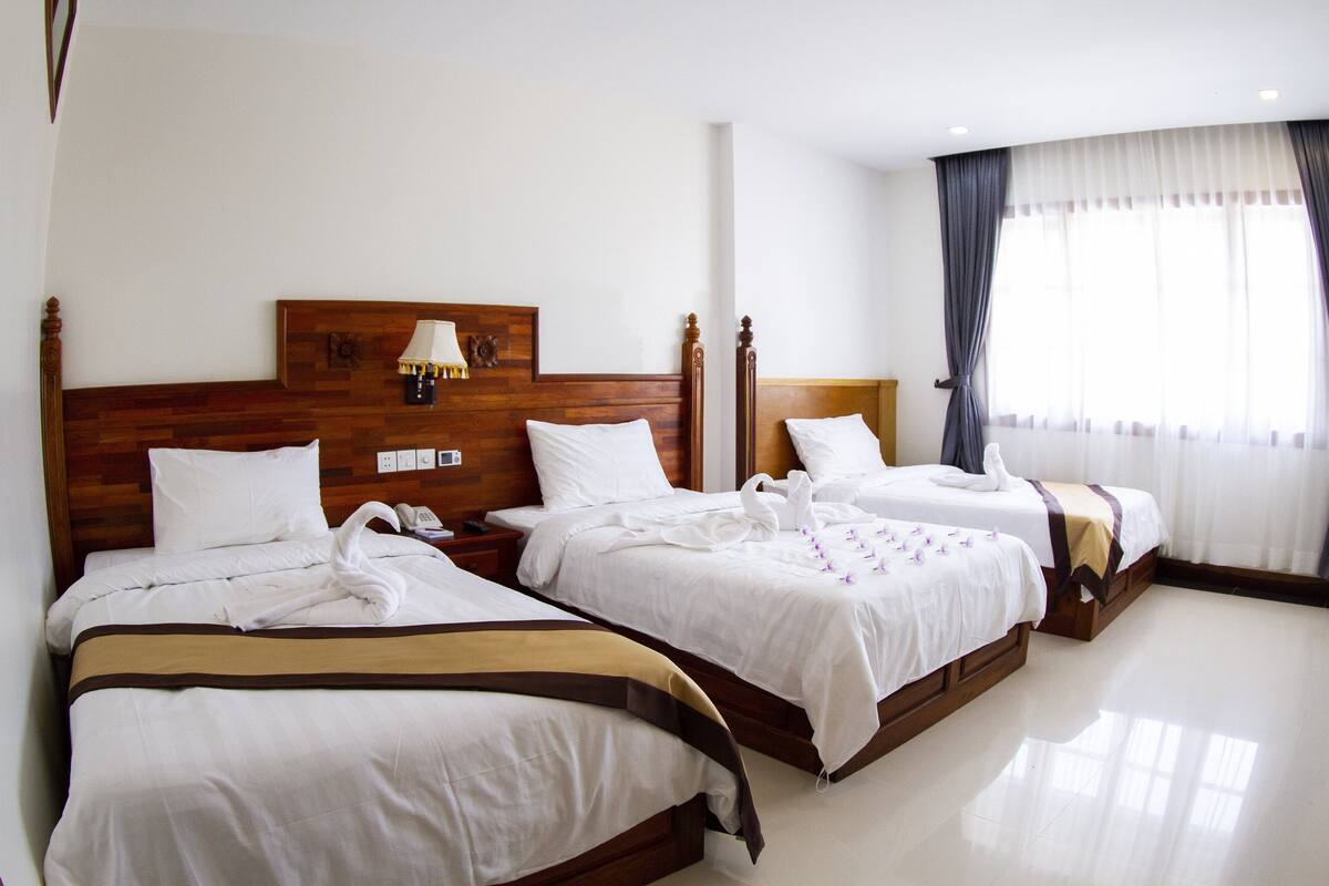 Habitación de hotel con tres camas, cabeceros de madera, ropa de cama blanca y una mesita de noche con lámpara.