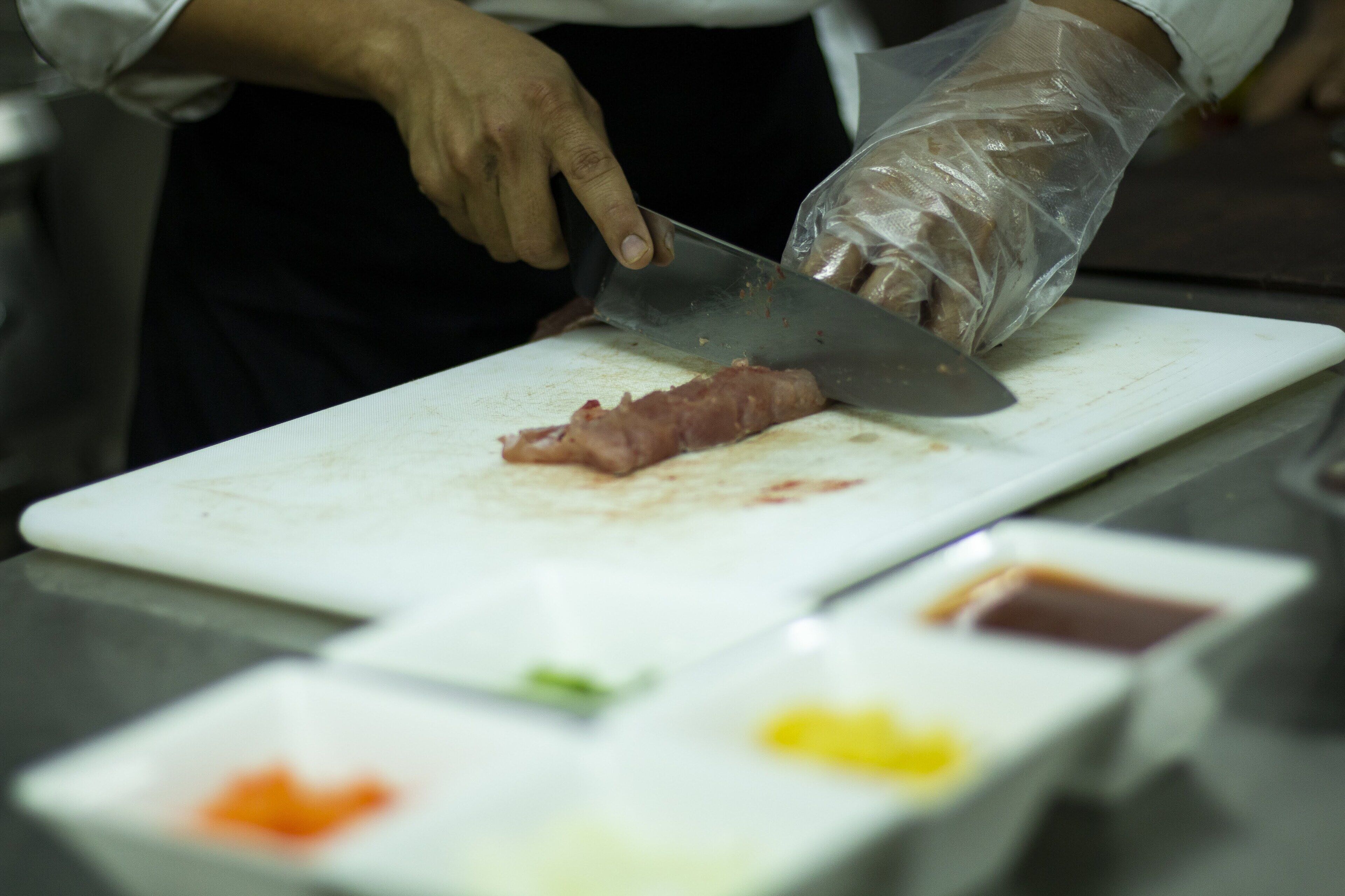 Una persona cortando carne sobre una tabla de cortar blanca con un cuchillo grande. Hay recipientes pequeños con salsas de colores diferentes frente a la tabla.