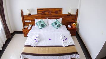 Una cama bien hecha con almohadones florales blancos y verdes, un cabecero de madera y una mesita de noche con una lámpara y una cesta de frutas.
