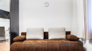 A brown leather sofa with two pillows against a white wall, a clock, and a window with curtains. - Modern Apartment in The Heart of Vienna 2 (Vienna)