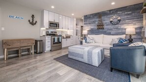 A modern living room with a sofa, ottoman, and a bench. There is a kitchen area with white cabinets and a microwave. The room has a gray wall with a 'BEACH' sign and a circular 'AT THE BEACH' sign. - Lakeside Serenity Villa with Private Patio (Bridgman)