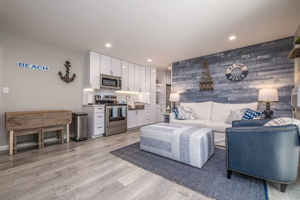 A modern living room with a sofa, ottoman, and a bench. There is a kitchen area with white cabinets and a microwave. The room has a gray wall with a 'BEACH' sign and a circular 'AT THE BEACH' sign.
