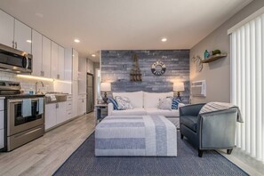 A modern kitchen with stainless steel appliances, a dining area with a sofa and lamps, and a bedroom with a bed and a nightstand.