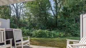A covered patio with white rocking chairs and a table, surrounded by lush greenery. - Lakeside Serenity Villa with Private Patio (Bridgman)