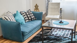 Ein Wohnzimmer mit einem petrolfarbenen Sofa, einem Glastisch, einem weißen Sessel und einem gemusterten Teppich.