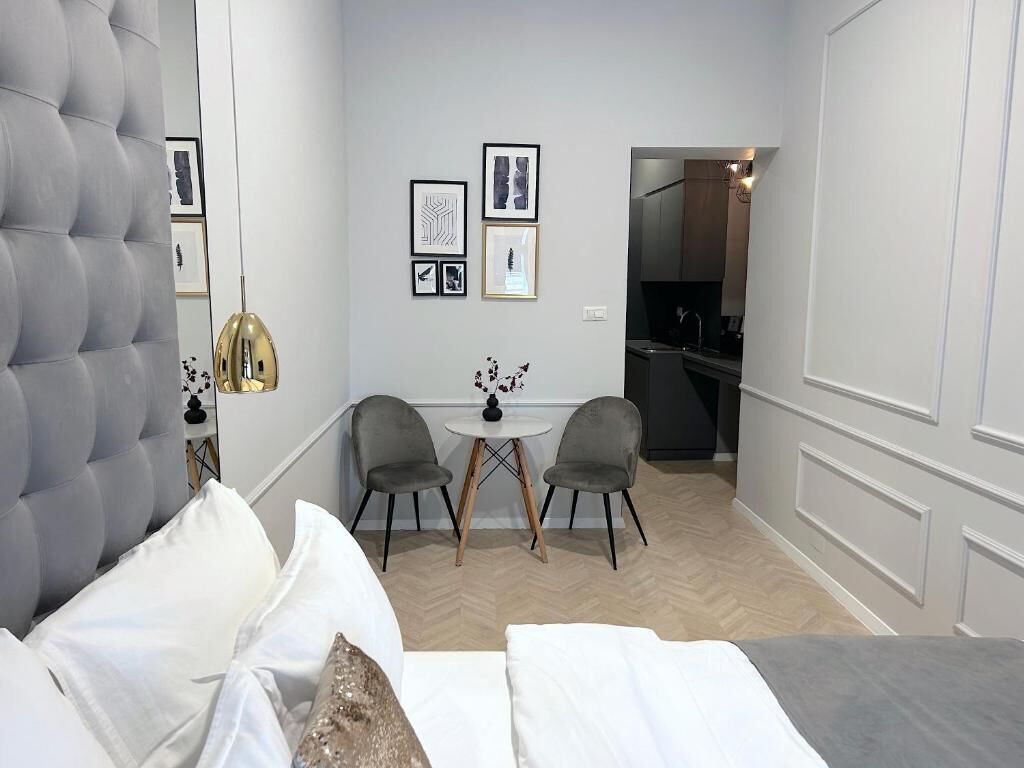 Una habitación de hotel compacta con cabecera capitonada, cama con ropa blanca, una pequeña mesa redonda con dos sillas y una kitchenette al fondo.