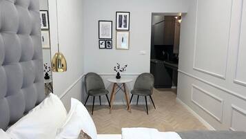 Una habitación de hotel compacta con cabecera capitonada, cama con ropa blanca, una pequeña mesa redonda con dos sillas y una kitchenette al fondo.