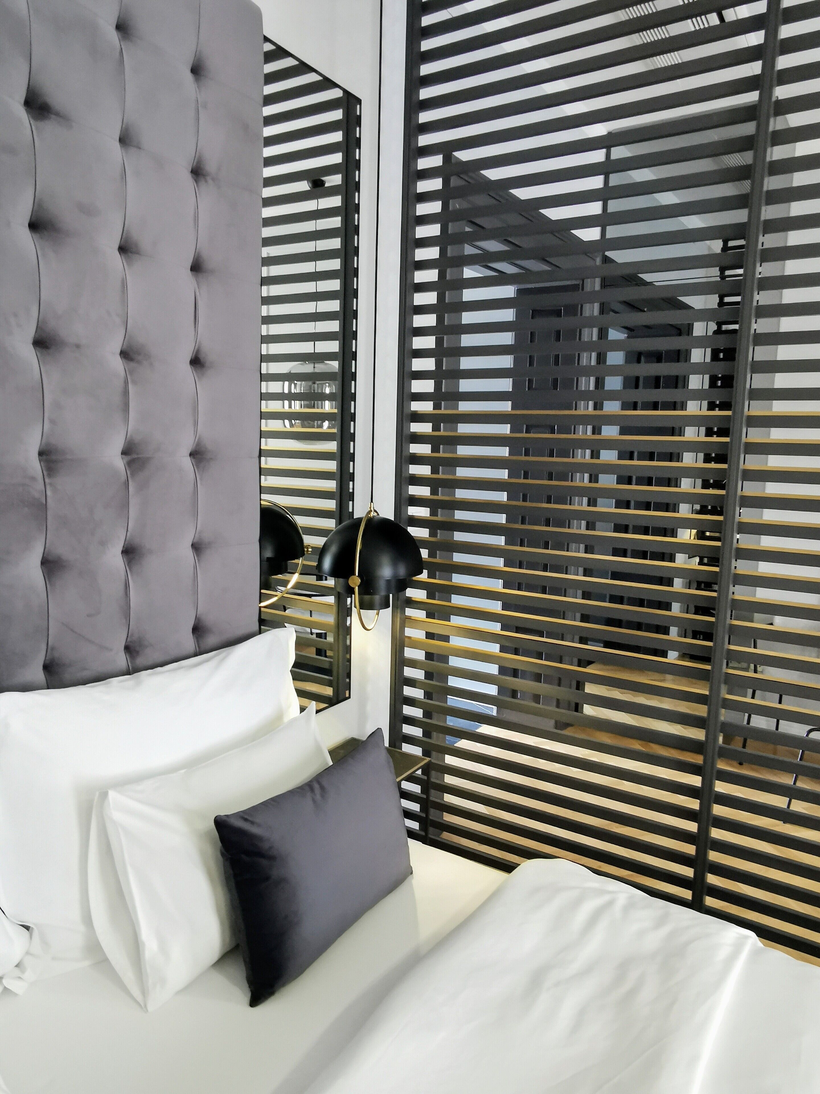 Une chambre à coucher moderne avec une tête de lit capitonnée, un lit avec des oreillers blancs et foncés, une lampe noire et une fenêtre avec des stores horizontaux.