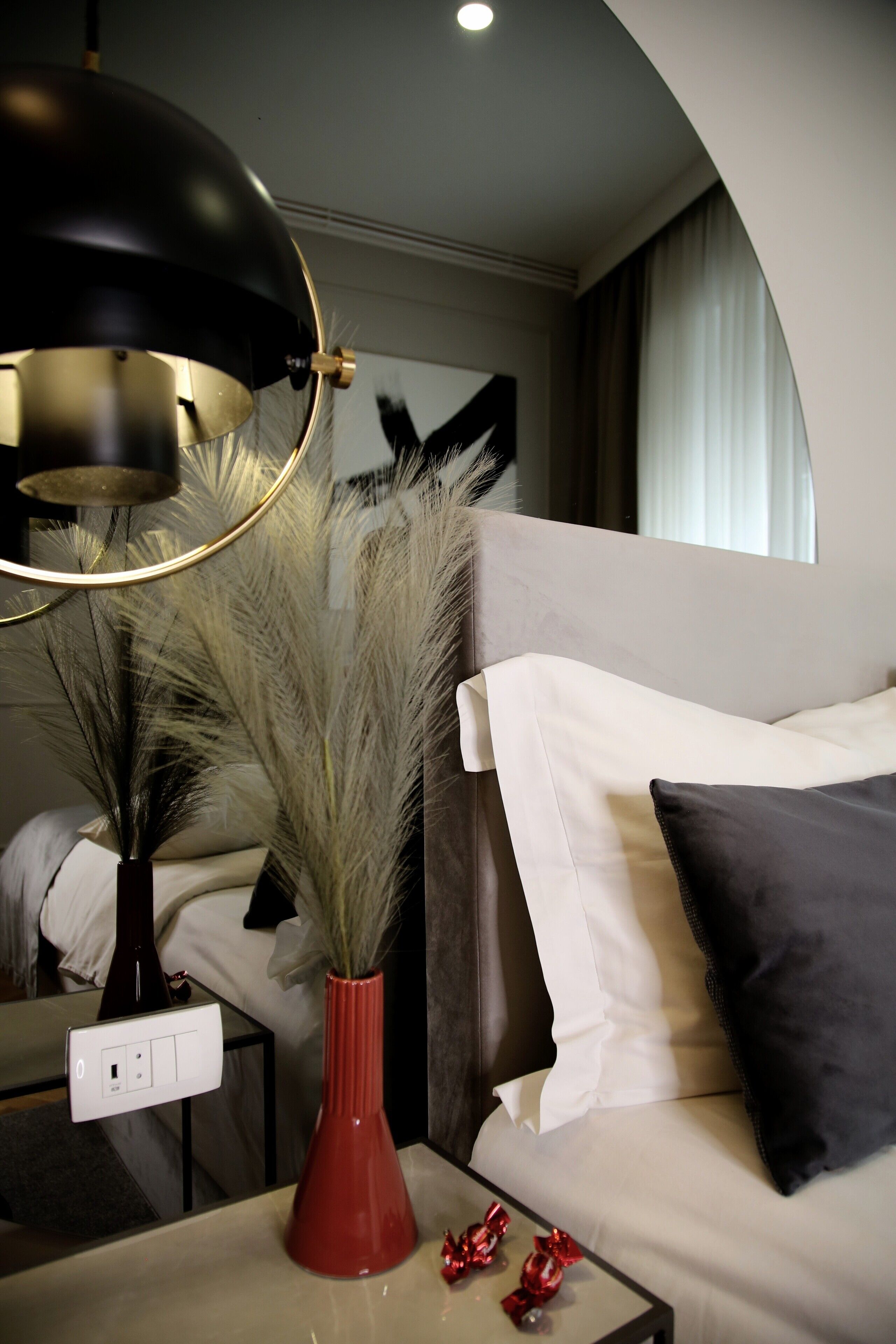 A modern hotel room with a sofa, a red vase with decorative plants, a black lamp, and a small table with a power outlet.