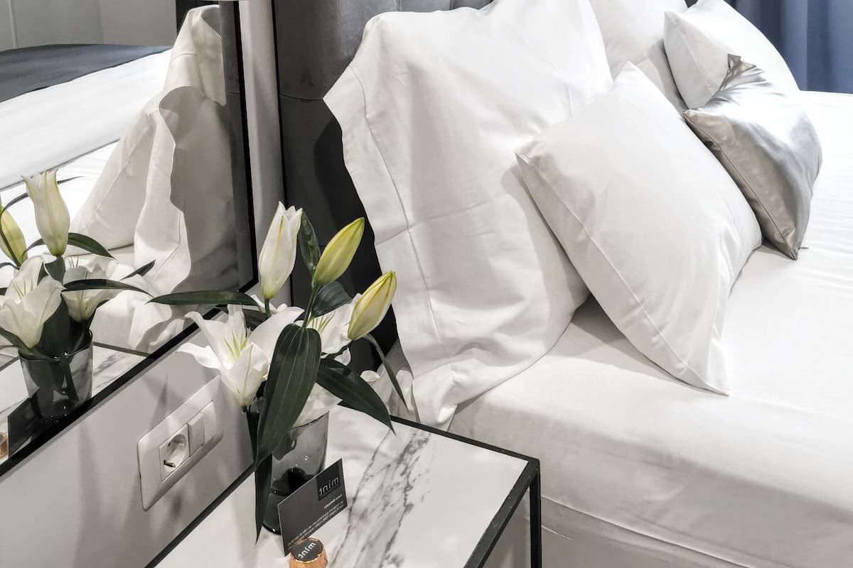 Une chambre d’hôtel moderne avec une tête de lit capitonnée, un grand miroir et une table de chevet agrémentée d’un vase de fleurs blanches.
