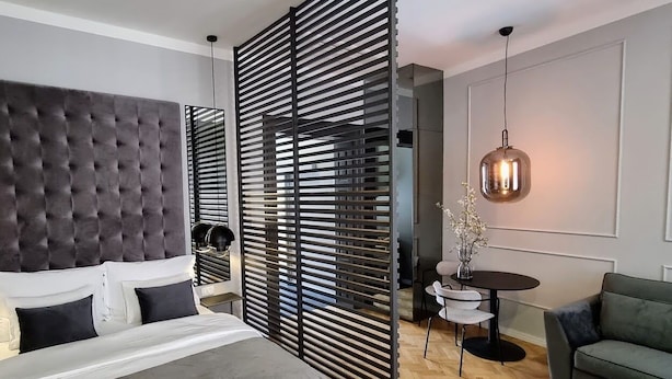 A modern bedroom with a tufted headboard, a large bed, a partition with horizontal slats, a small round table with chairs, a hanging pendant light, and a green sofa. - Mint Boutique Studio Apartments  (Zagreb)