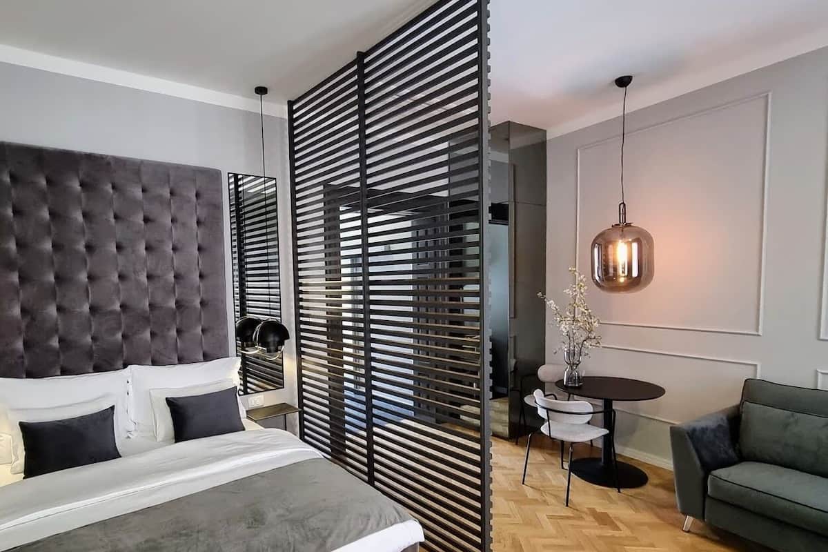 Une chambre moderne avec une tête de lit capitonnée, un grand lit, un séparateur composé de lattes horizontales, une petite table ronde avec des chaises, une suspension et un canapé vert.