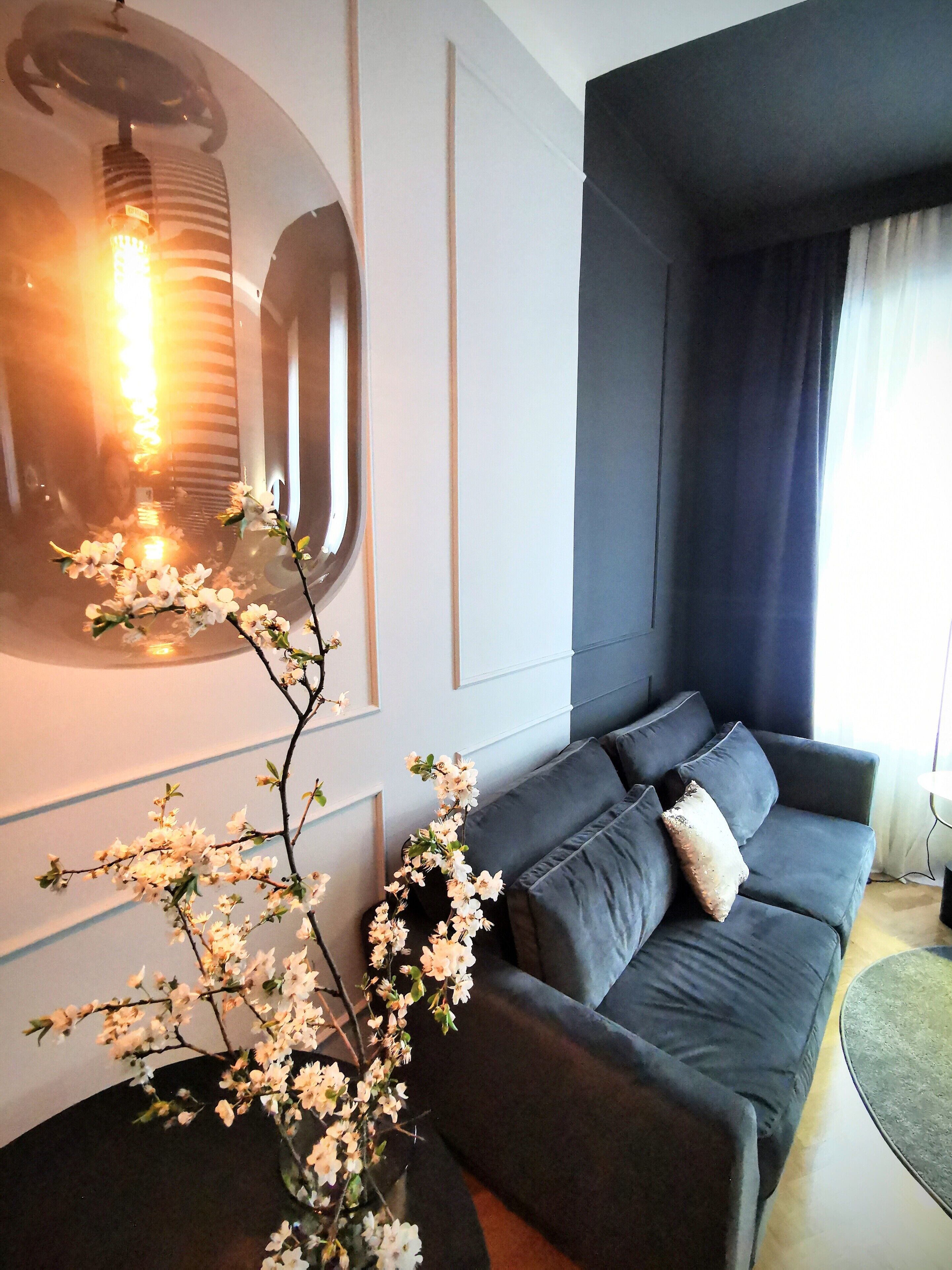 Una sala de estar moderna con un sofá gris oscuro, un florero con flores blancas y una lámpara fijada a la pared.