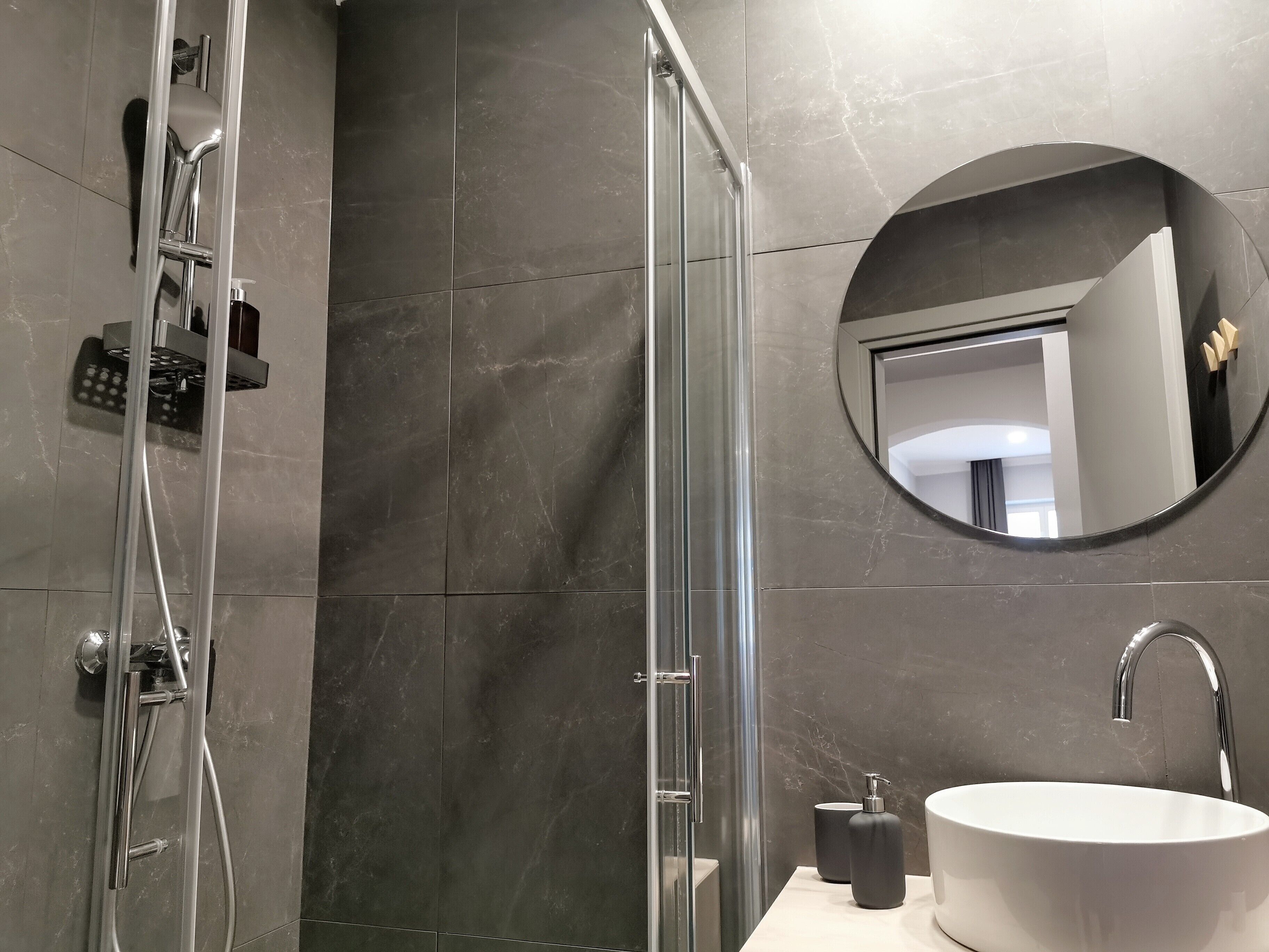 A modern bathroom with a glass-enclosed shower, a round mirror, a white sink, and a handheld showerhead.