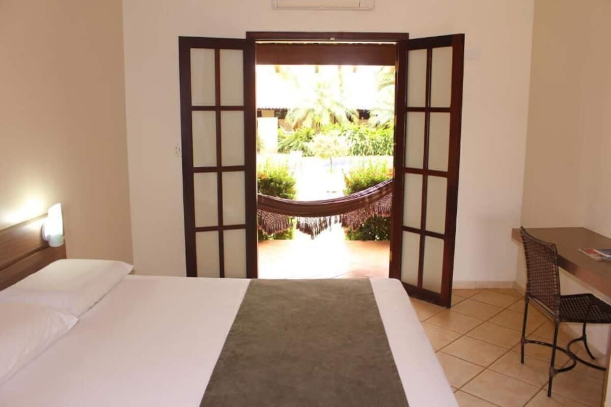 A hotel room with a bed, a desk, and a view of a hammock and greenery outside.