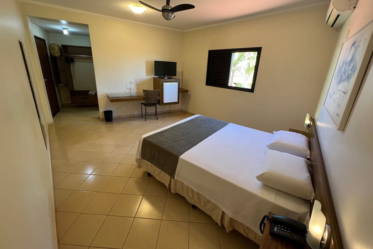 A hotel room with a bed, a desk with a computer, a chair, a window, and a ceiling fan.