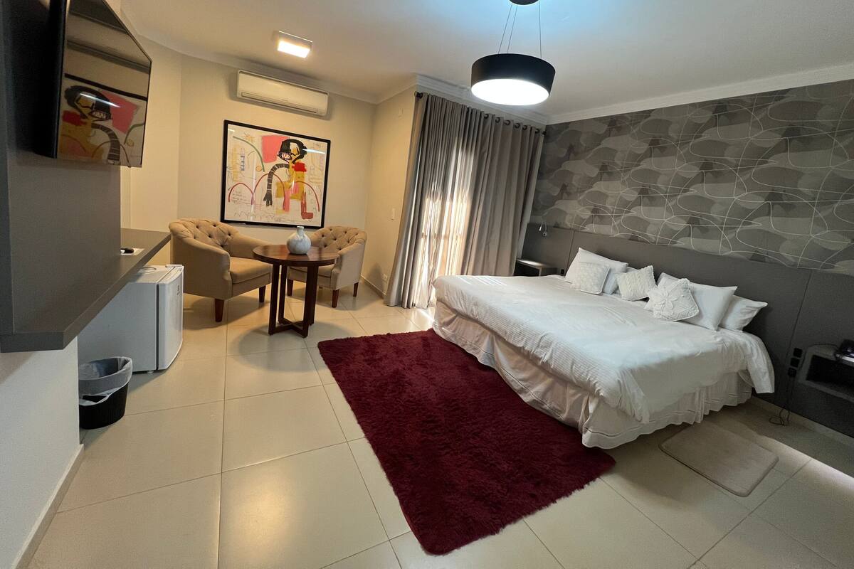 A hotel room with a bed, a desk, a chair, a red rug, and a painting on the wall.
