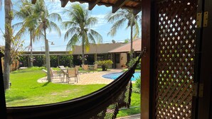 Una hamaca bajo una pérgola de madera con vista a una piscina, palmeras y un área de patio.