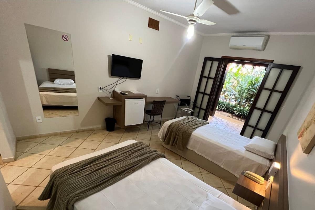 A hotel room with two beds, a desk with a computer, a TV, and a window with a view of greenery.