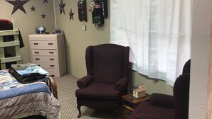 Een slaapkamer met een bed, een kaptafel, twee stoelen, een raam met gordijnen en wanddecoraties, waaronder sterren en een klok.