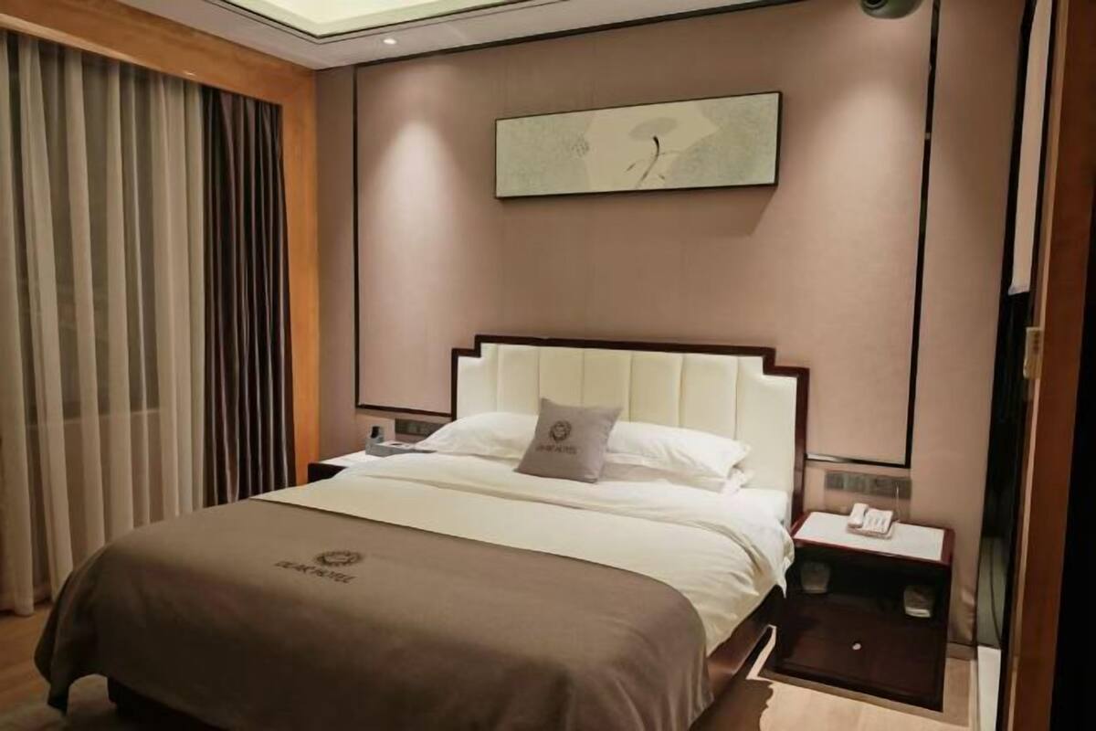 A hotel room with a bed, a nightstand, a clock on the wall, and a window with curtains.